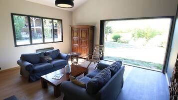 Un salon spacieux avec deux canapés, une table basse et un meuble en bois. De grandes fenêtres offrent une vue sur l’extérieur.