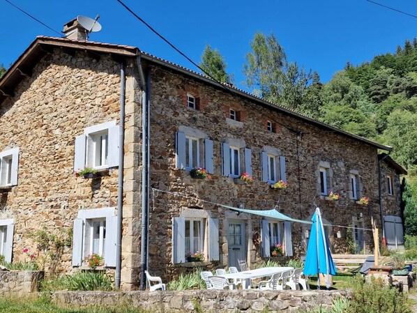 Un bâtiment en pierre doté de nombreuses fenêtres, dont certaines sont équipées de volets, et d’un espace extérieur avec des sièges et un parasol bleu.