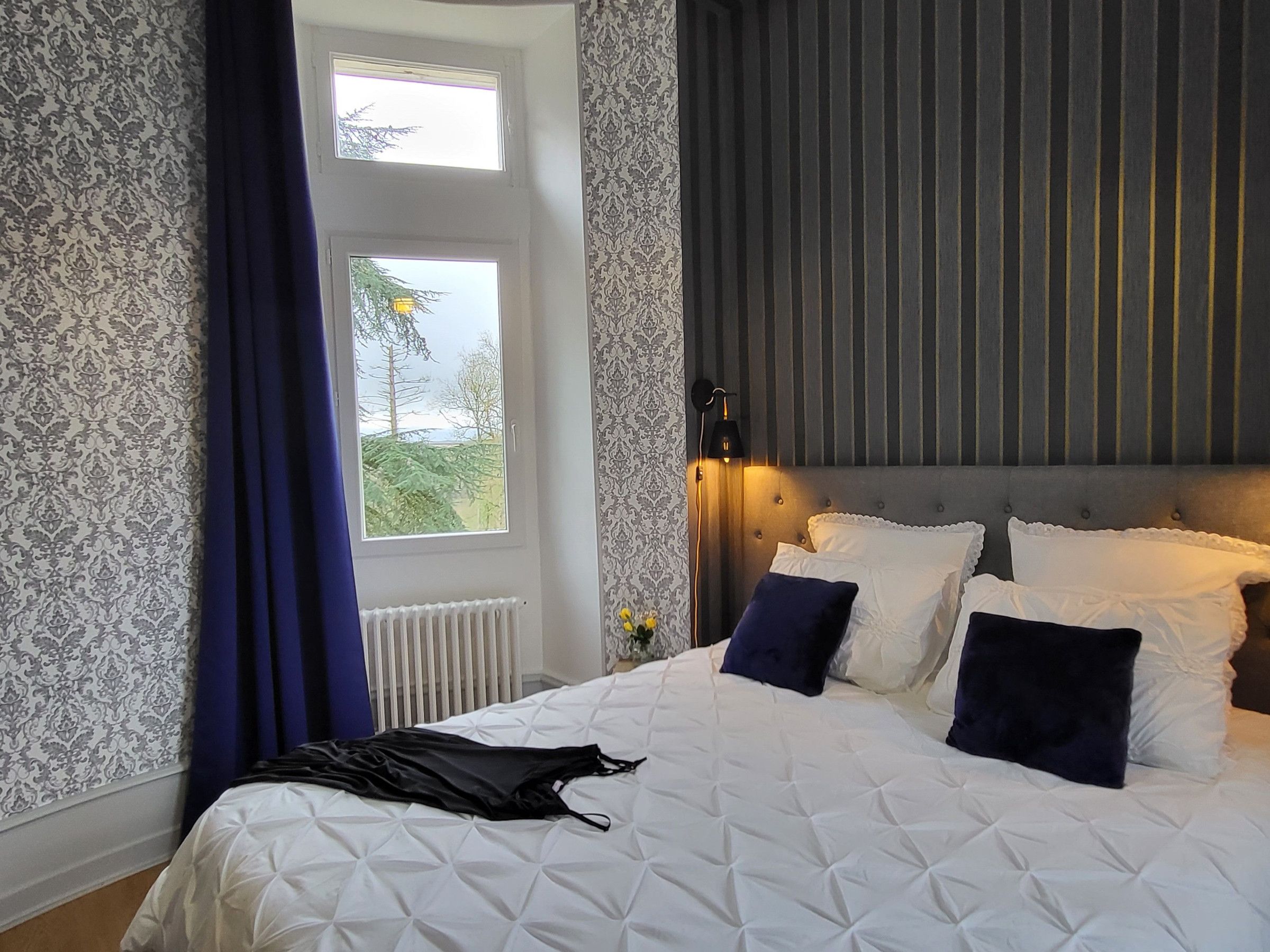 Une chambre avec un grand lit, une fenêtre avec des rideaux, un radiateur et une lampe fixée au mur.