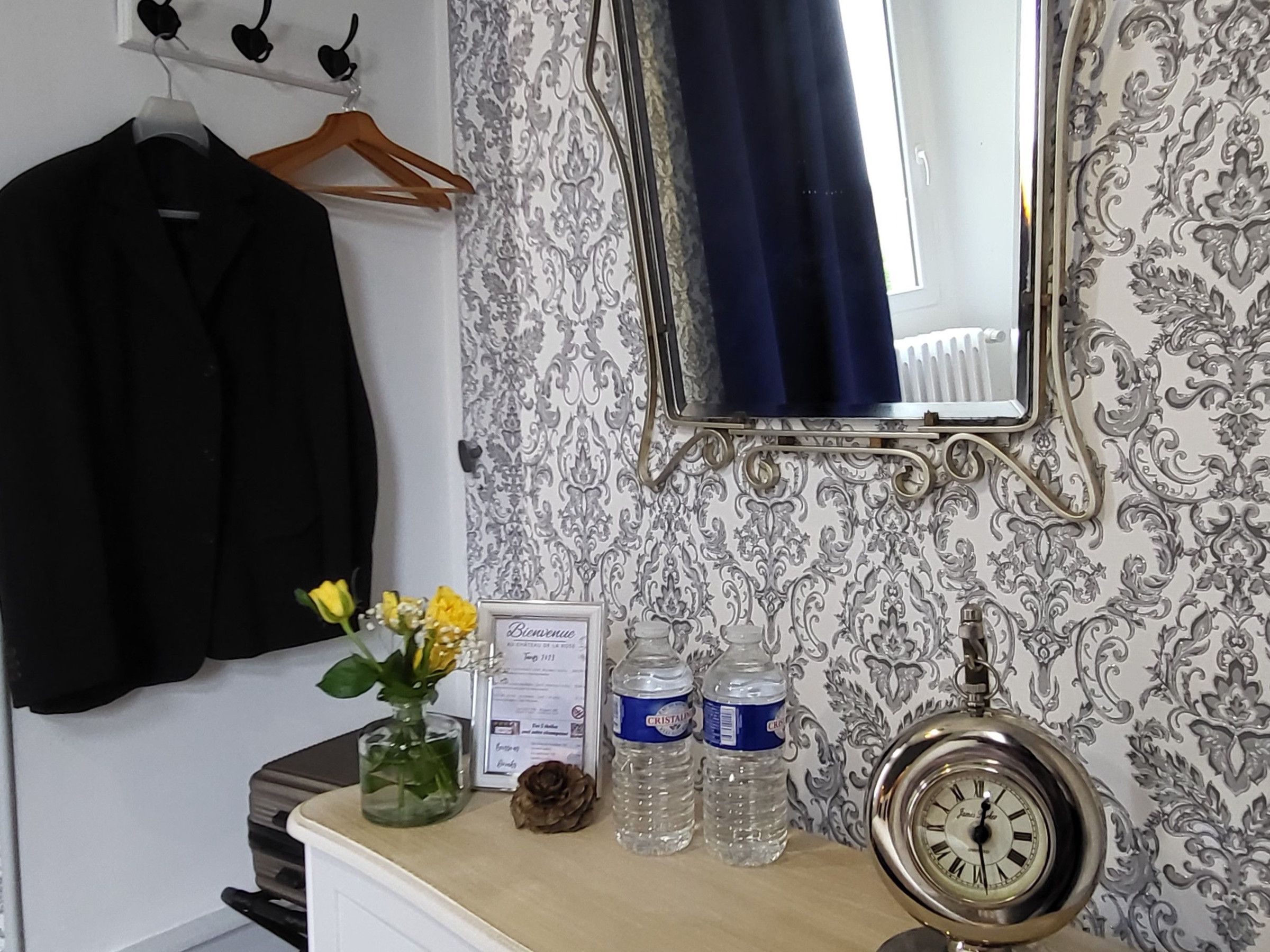 A hotel room with a mirror, a clock, a vase with flowers, a water bottle, and a black jacket hanging on a rack.