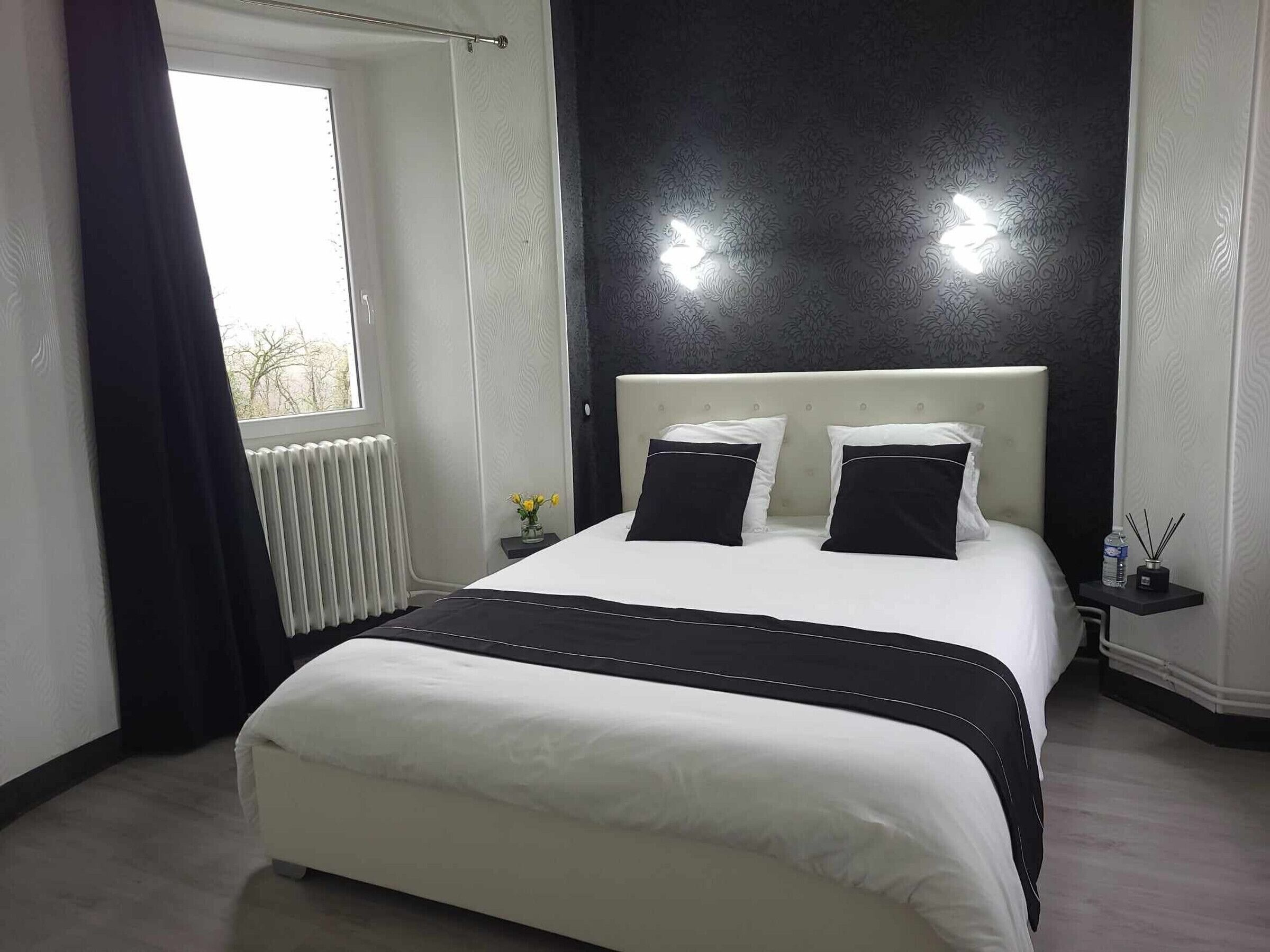 A modern bedroom with a large bed, a radiator, a window with curtains, and a nightstand with a water bottle and a candle holder.