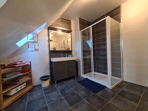 A modern bathroom with a shower, dark tiled floor, and a wooden shelf with towels. - Cozy room with sitting area and breakfast included (Châteauroux)