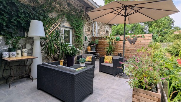 An outdoor patio area with wicker furniture, a large umbrella, potted plants, and a stone wall.