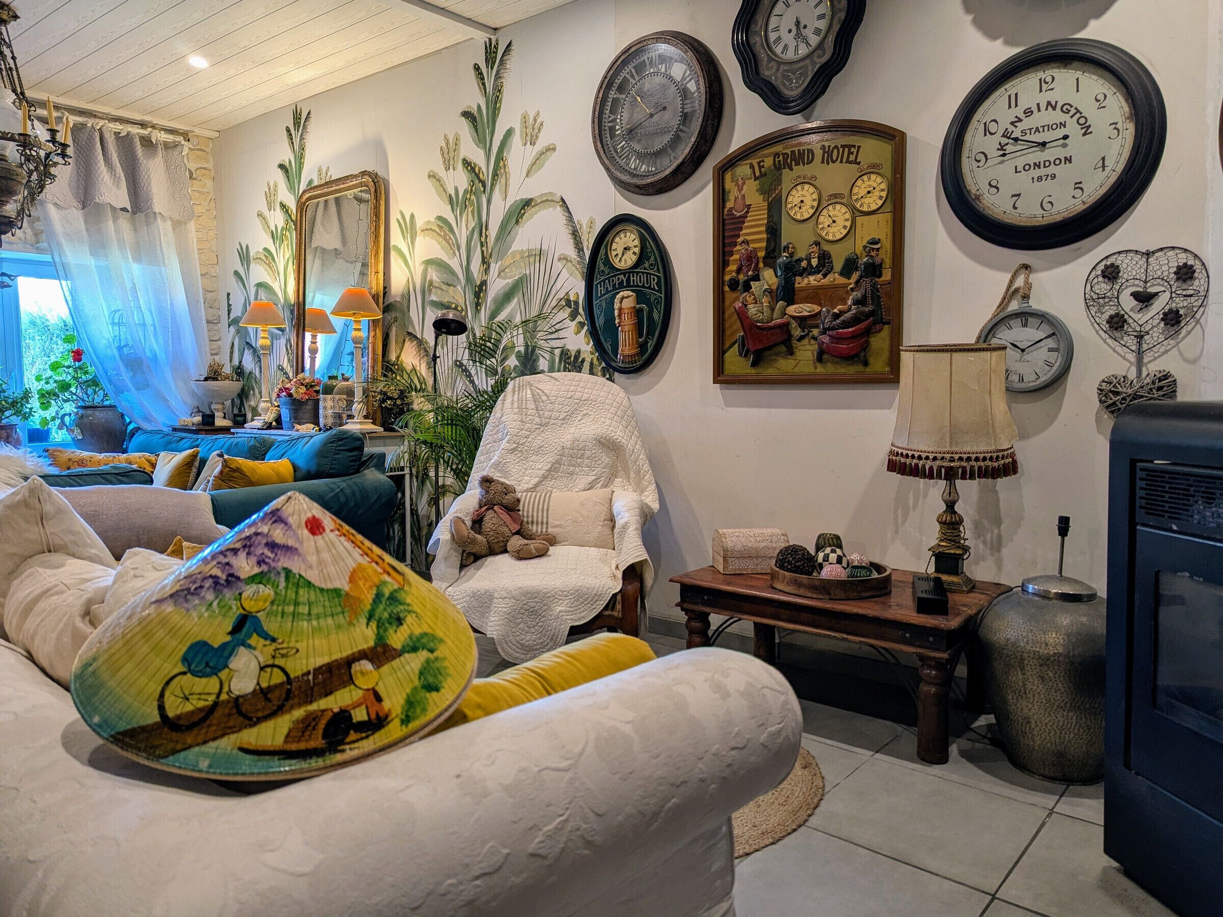 Una sala de estar acogedora con un sofá, un sillón, una mesa con lámpara y una chimenea. Hay varios relojes en la pared y un cuadro que representa una escena de hotel.