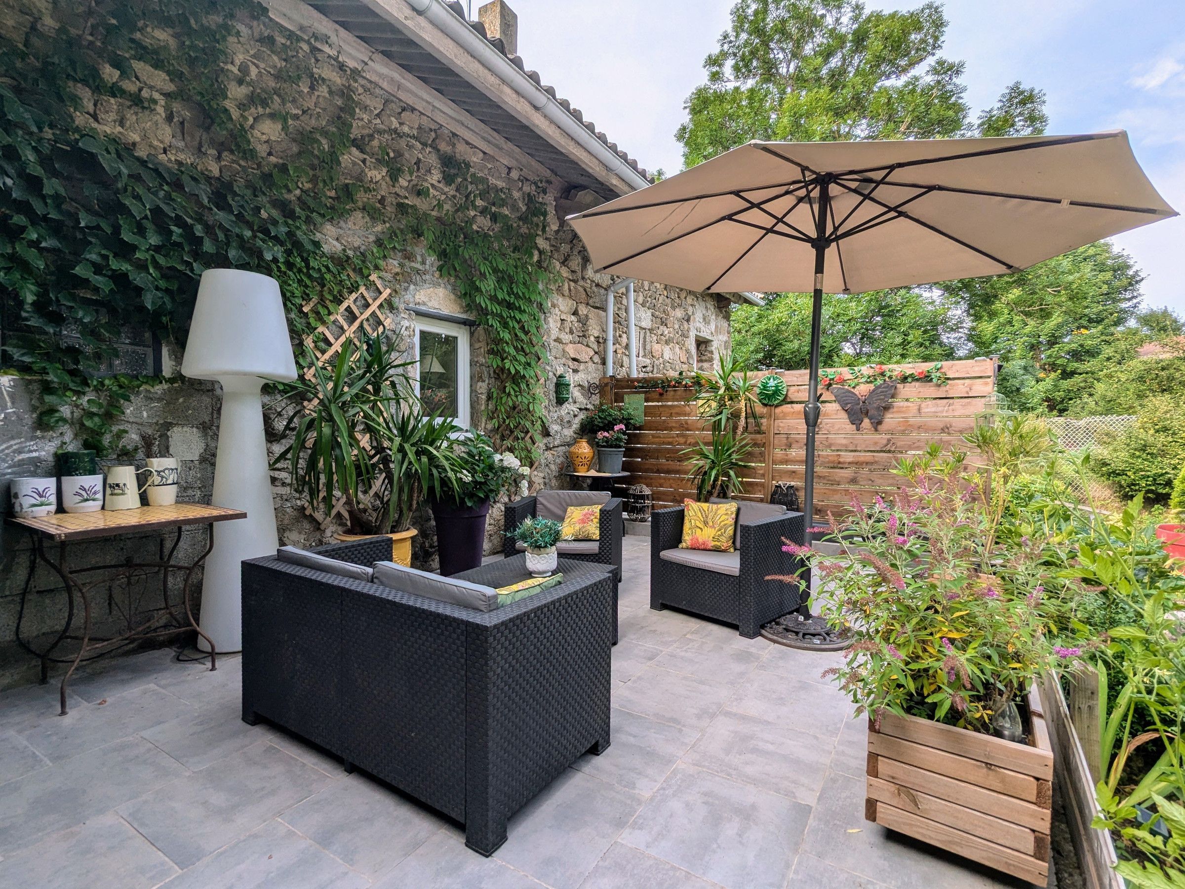 Un patio al aire libre con mobiliario de mimbre, un paraguas grande, plantas en macetas y una pared de piedra.
