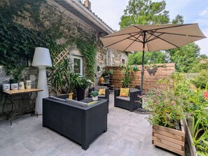 Een terras buiten met rieten meubels, een grote parasol, potplanten en een stenen muur.
