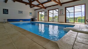 Une piscine intérieure dotée d’un sol carrelé, de poutres en bois au plafond et de grandes fenêtres offrant une vue sur l’extérieur.
