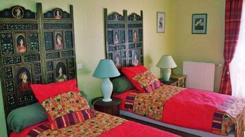 Ein Zimmer mit zwei Betten, jeweils mit einem blumigen Überwurf und roten Kissen. Zwei Nachttische mit Lampen, und ein dekorativer Kopfteil mit eingekachelten Bildern.