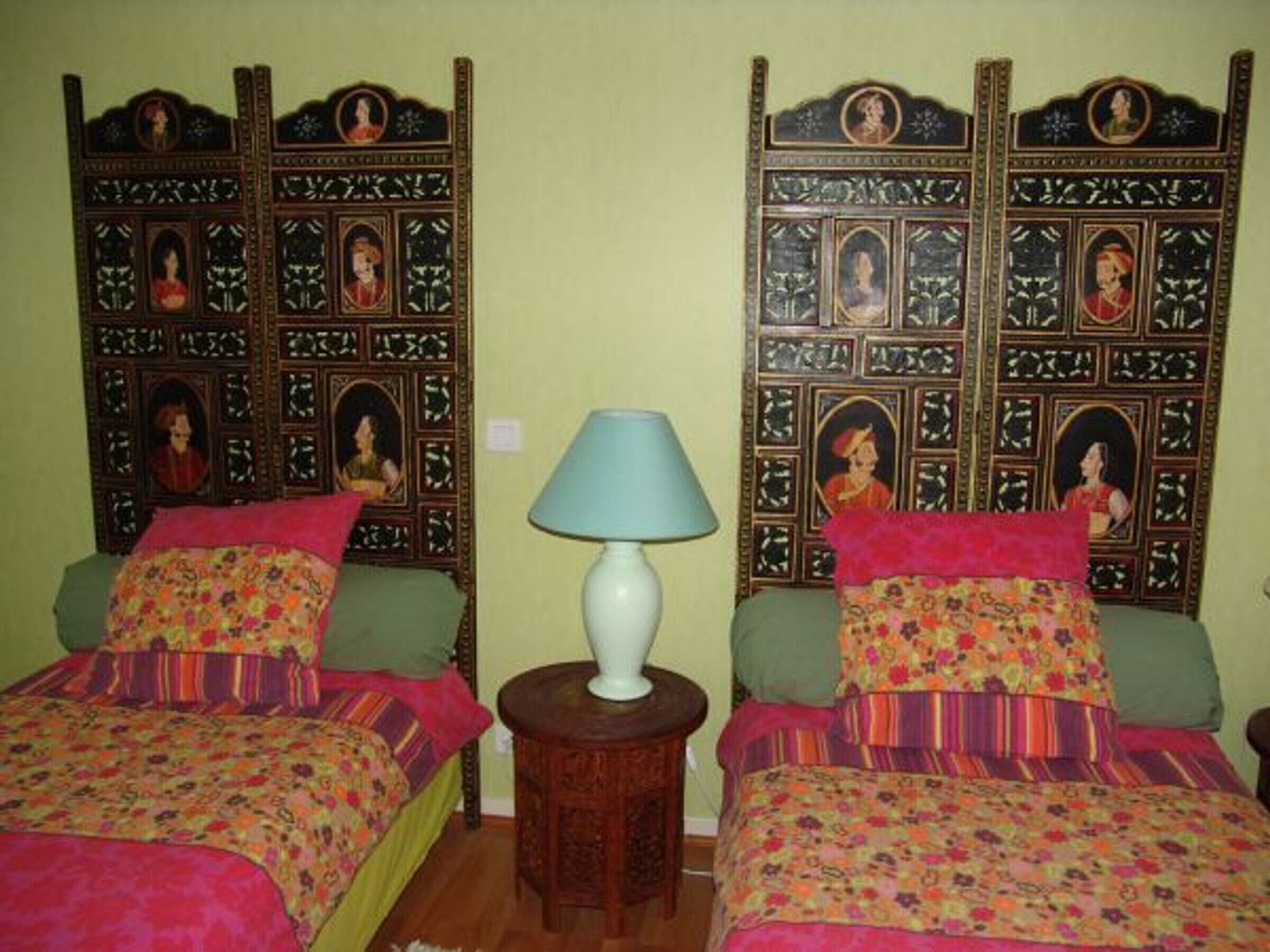 A room with two beds, each with a floral bedspread and a green pillow. There is a white lamp on a wooden side table between the beds.