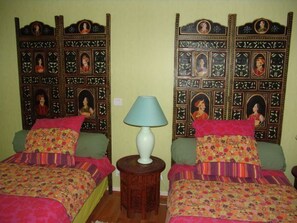 Ein Zimmer mit zwei Betten, jedes mit einer blumigen Tagesdecke und einem grünen Kissen. Zwischen den Betten steht ein Holznachttisch mit einer weißen Lampe.