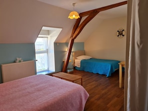 A cozy bedroom with a sloped ceiling, a wooden beam, a window, a radiator, a bed with a blue bedspread, and a bedside table.
