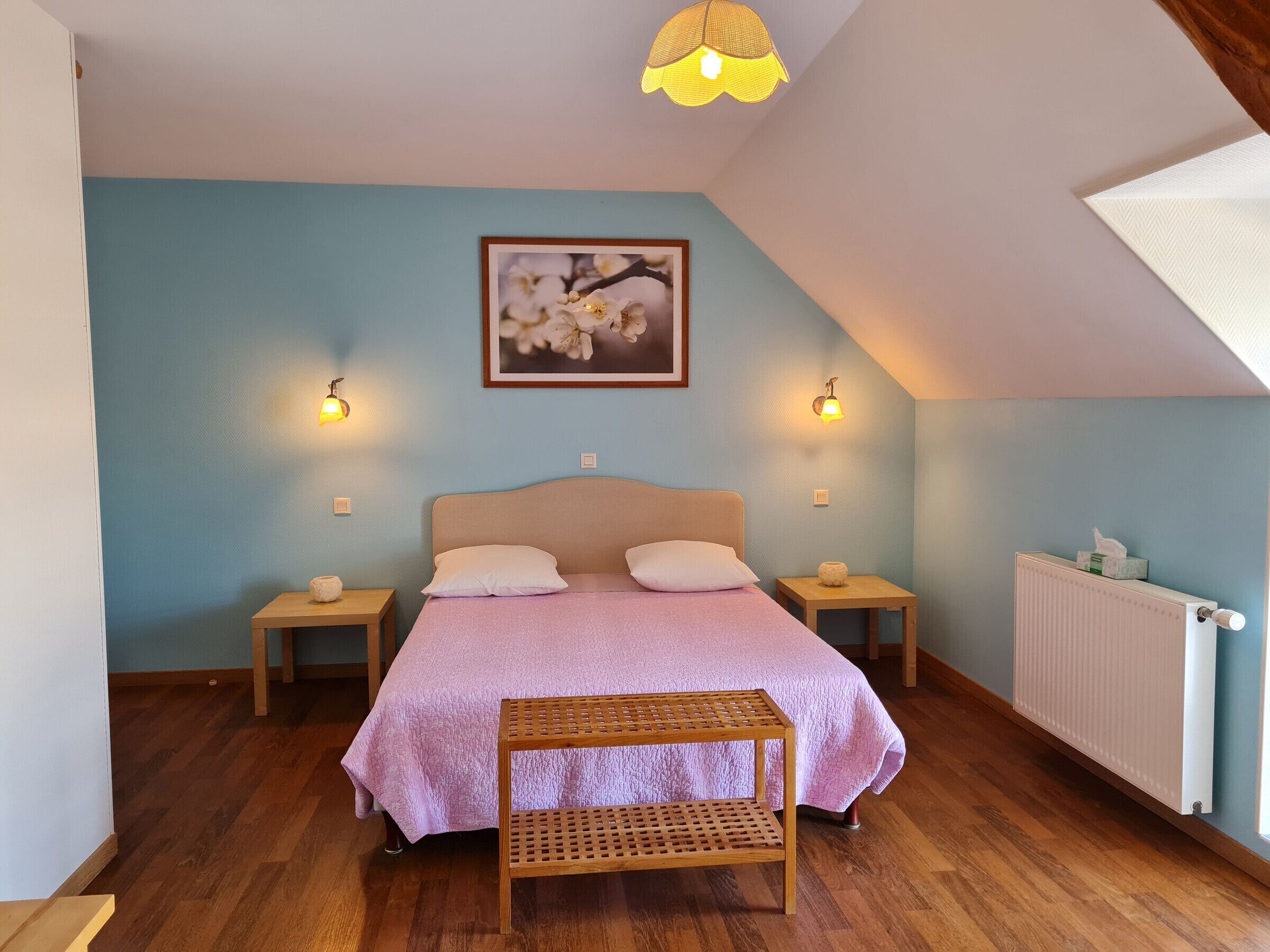 A bedroom with a sloped ceiling, a bed with a pink quilt, two bedside tables with lamps, a wooden bench, a radiator, and a framed picture on the wall.