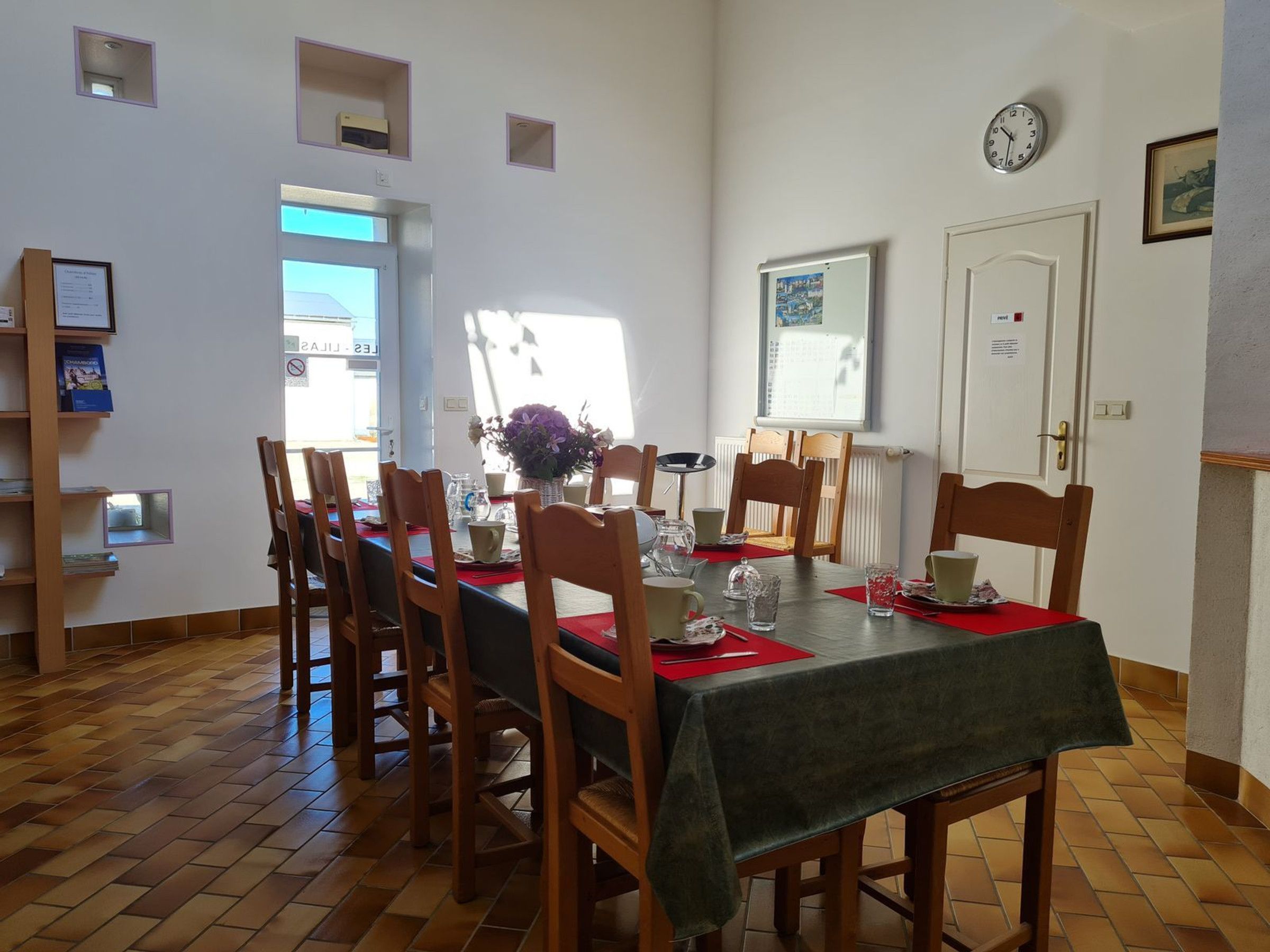 A dining area with a table set for six, wooden chairs, a clock on the wall, and a door with a sign.