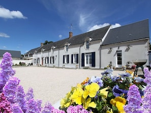 A row of white buildings with dark shutters, a gravel courtyard, and a variety of flowers in the foreground.