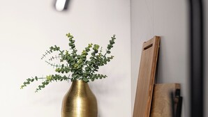A brass vase with green foliage on a grey countertop, a wooden board leaning against a wall, and a black curved object. - RentPlanet - Studio Kępna (Warsaw)