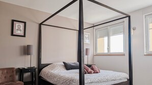 Un lit à baldaquin avec une couvre-lit à motifs floraux, une chaise, une table de chevet avec des lampes et une fenêtre avec des stores.