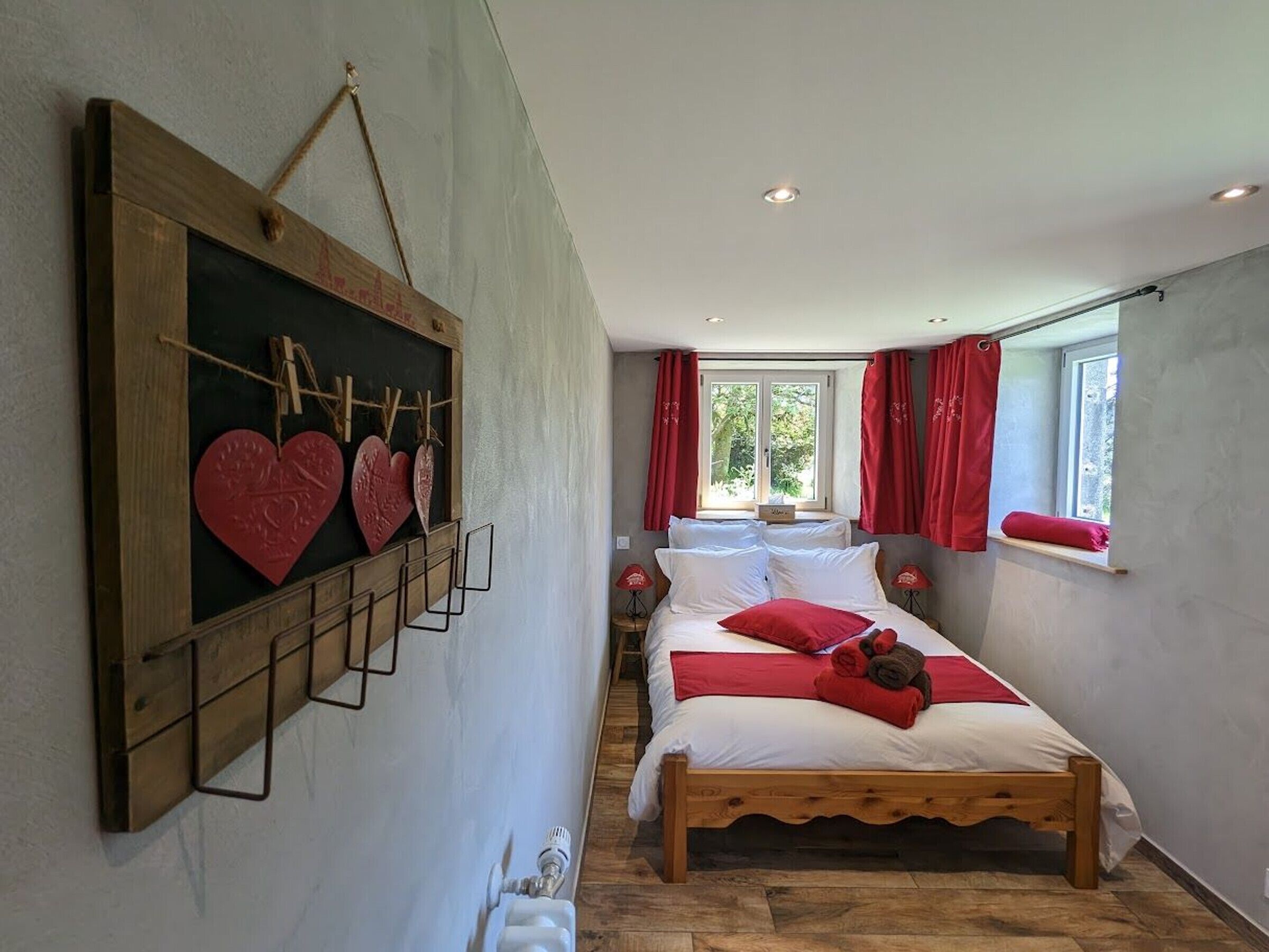 Un dormitorio con cama de madera, estante de pared con adornos con forma de corazón y ventana con cortinas rojas.
