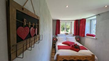 Un dormitorio con cama de madera, estante de pared con adornos con forma de corazón y ventana con cortinas rojas.