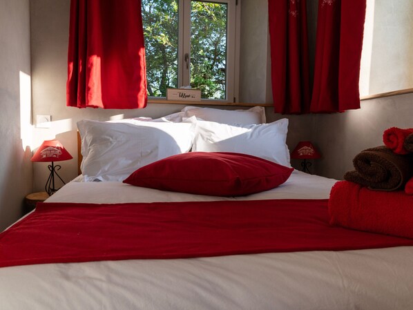 Ein ordentlich bezogenes Bett mit weißer und roter Bettwäsche, einem roten Kissen und einer roten Decke. Ein Fenster mit roten Vorhängen, ein Nachttisch mit einer Lampe und ein Schild mit der Aufschrift 'Tissue Box'.