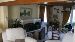 Una sala de estar con televisor, dos sillones blancos, una mesa de comedor de madera con camino de mesa verde, y una cocina al fondo.