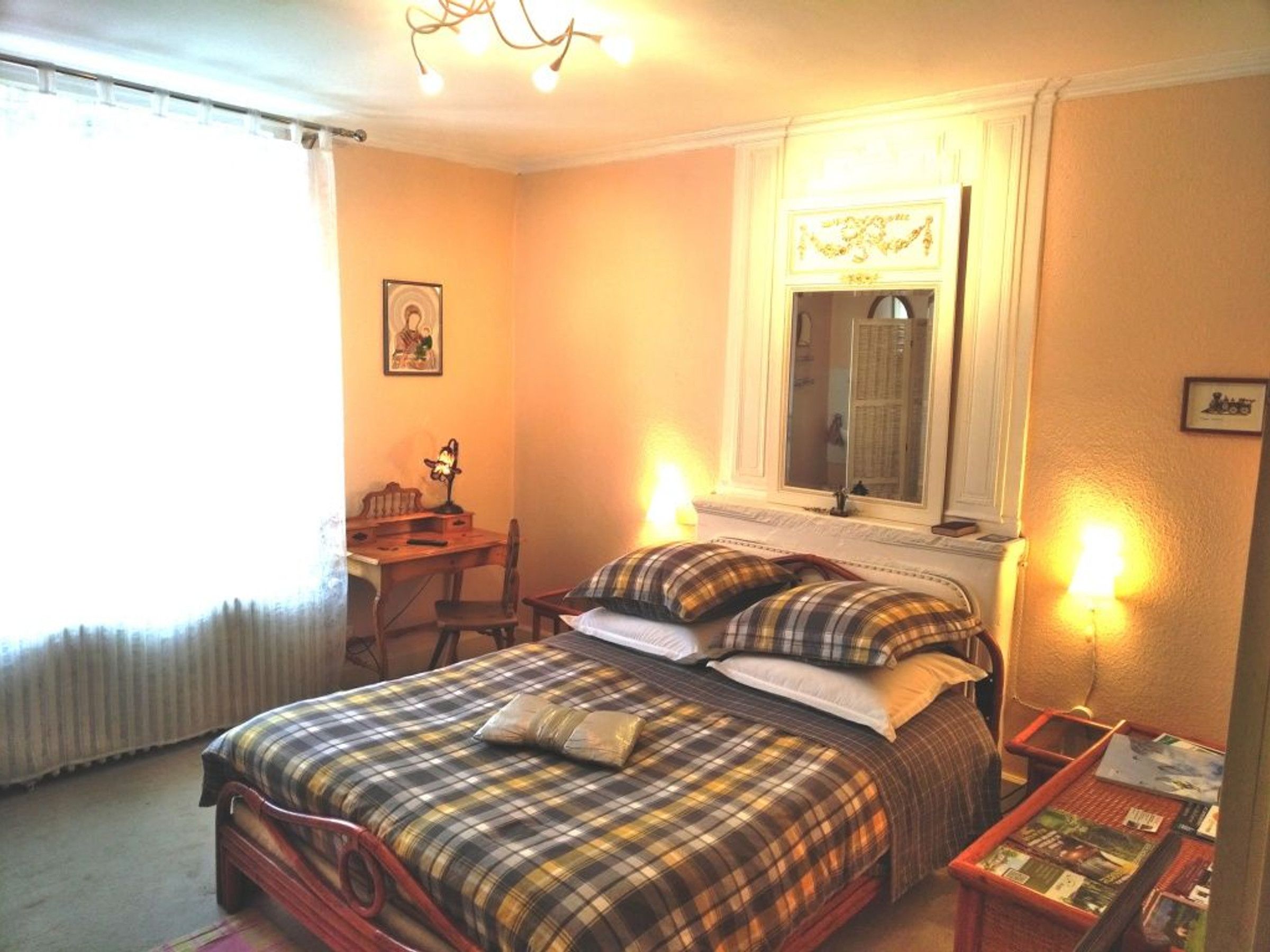 Une chambre à coucher avec un lit, un bureau et une chaise. Il y a une fenêtre avec des rideaux, un miroir au-dessus du lit et une lampe sur le bureau.
