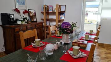 Uma sala de jantar com uma mesa posta para quatro pessoas, uma cafeteira, uma estante de livros e uma porta que dá para o exterior.