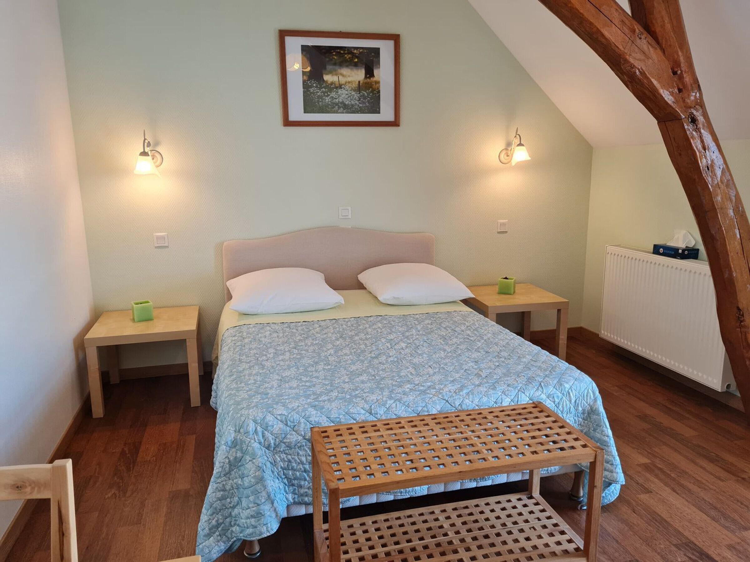A bedroom with a bed, two bedside tables, a nightstand, a wooden bench, a framed picture on the wall, and a radiator.