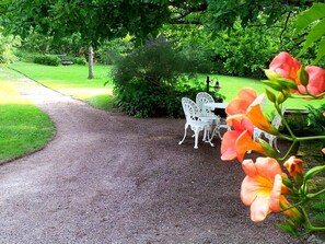 Un chemin de gravier mène à un ensemble de table et de chaises en métal blanc, placé sous un arbre.