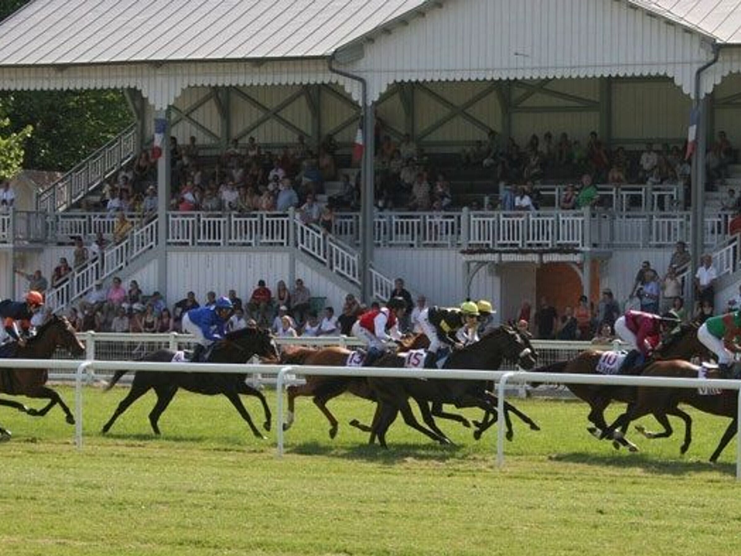 A horse race with jockeys on horses, a crowd of spectators, and a covered spectator stand.