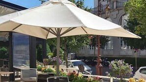 Un espace extérieur aménagé avec des tables et des chaises, protégé par un grand parasol, entouré de fleurs et offrant une vue sur la rue.