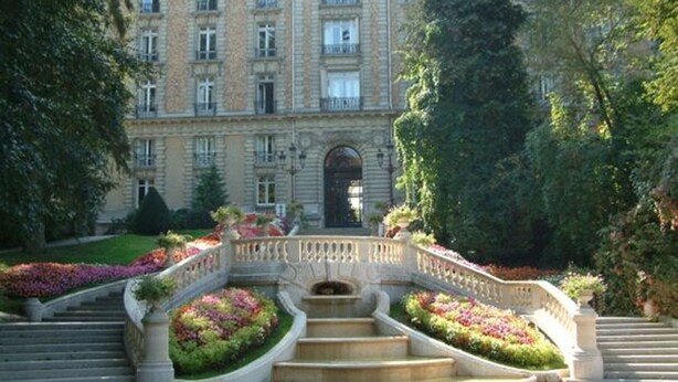 Un imposant bâtiment doté d’un escalier central, entouré de jardins luxuriants et agrémenté d’un pont décoratif.