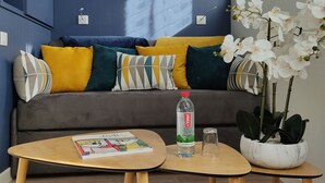 Un salon moderne avec un canapé, des coussins, une petite table sur laquelle se trouve un magazine, une bouteille d’eau et une plante en pot.
