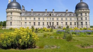 Un edificio grande e histórico con dos torres abovedadas destacadas, una fachada simétrica y un jardín bien cuidado con diversas plantas.
