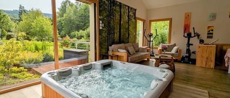 Un spa moderno con bañera de hidromasaje, suelo de madera, un sofá y vistas a un jardín exuberante.
