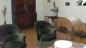 A room with a wooden cabinet, two armchairs, a table with a cake, and a window with plants.