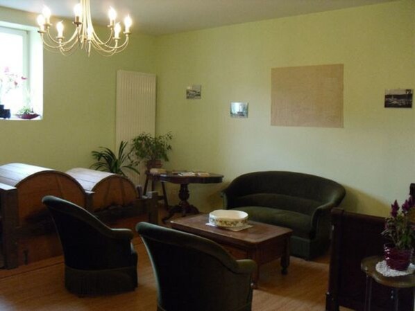 Una sala de estar acogedora con una mesa de centro de madera, un sillón verde y un sillón oscuro. Se observa una araña colgando del techo y una ventana que ofrece vistas al exterior.