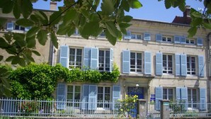 Un bâtiment à deux étages avec des volets bleus, une entrée fermée par un portail et un petit jardin agrémenté de fleurs et d’arbustes.