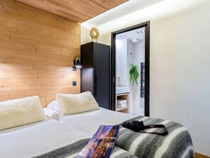 A modern hotel room with a wooden ceiling, a bed with white bedding, a black wardrobe, a bathroom visible through an open door, and a camera on the bed.