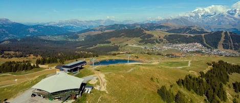 Ein modernes Gebäude mit Flachdach und großem Parkplatz, umgeben von einer gebirgigen Landschaft und einer kleinen Stadt in der Ferne.