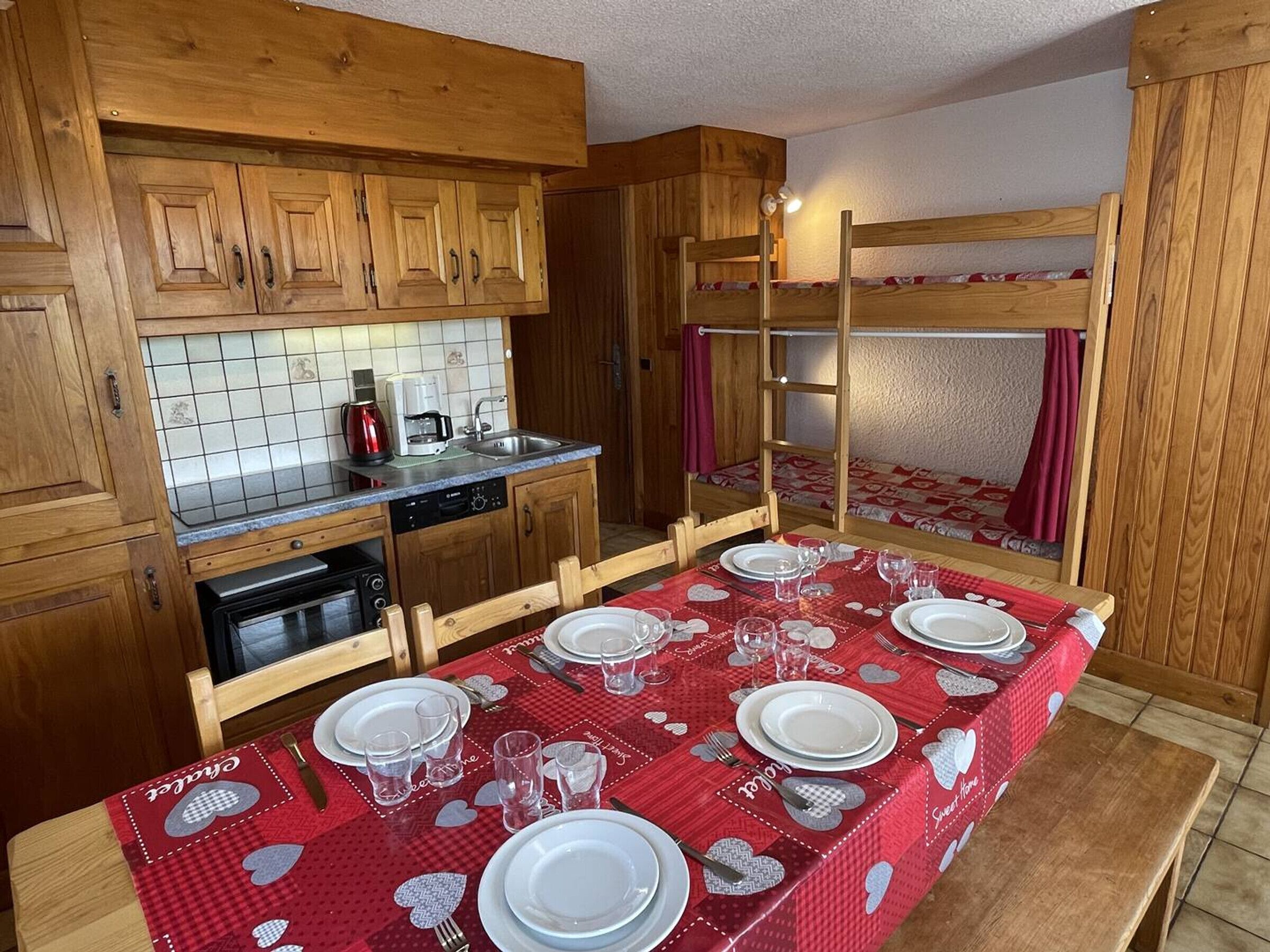 Ett kompakt kök med träskåp, kakel ovanför arbetsbänken och en matplats med plats för fyra personer, där bordet är dukat med en röd duk.