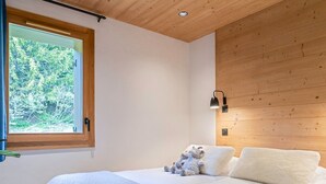 A bedroom with two beds, a window with a view of greenery, a wooden ceiling, and a wall-mounted lamp.