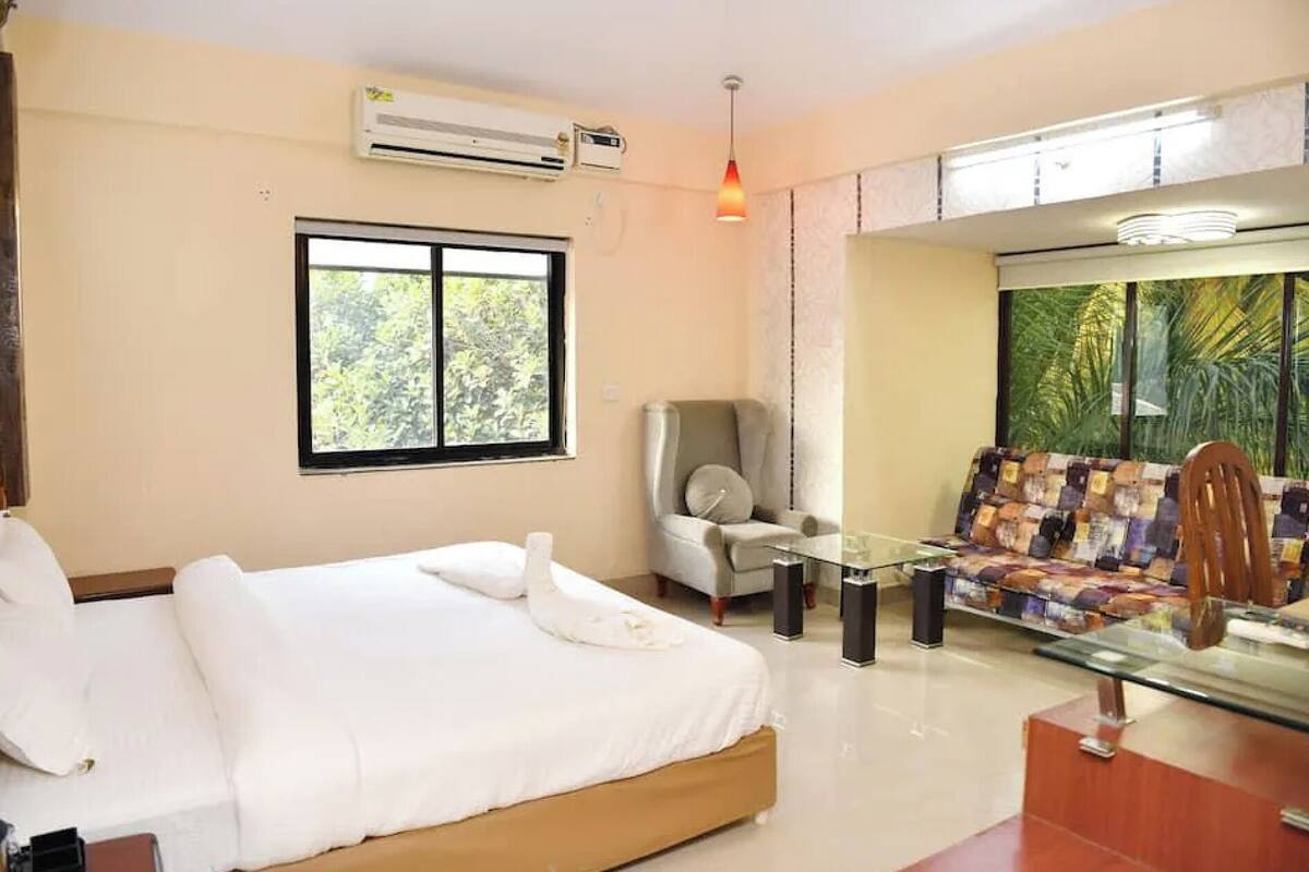 A hotel room with a bed, a chair, a sofa, a glass table, and a view of greenery outside.