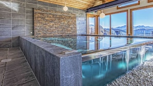 A modern spa area with a glass-enclosed hot tub, stone walls, wooden ceiling, and a view of mountains.