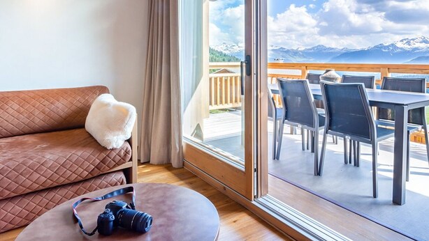 A room with a wooden ceiling, a brown sofa with a white throw pillow, a round table with a camera, and a balcony with outdoor seating and a mountain view.
