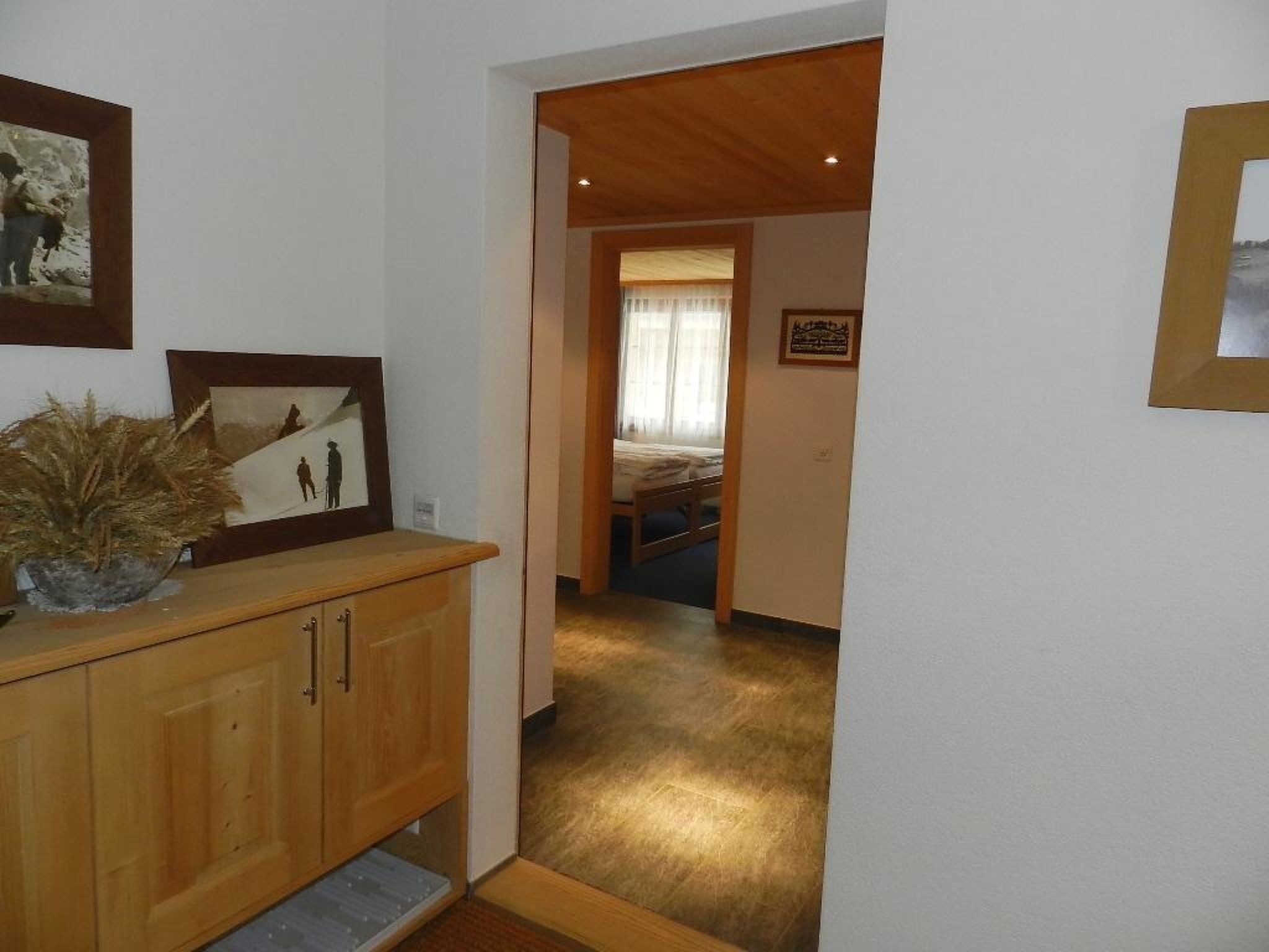 A hallway leading to a bedroom with a bed and a nightstand. There is a wooden cabinet with a plant on top and framed pictures on the wall.