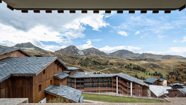 Un edifici de fusta amb teulada inclinada, envoltat de muntanyes i un cel serè.
