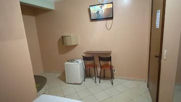 A hotel room with a bed, a small table with chairs, a refrigerator, a television mounted on the wall, and a door.