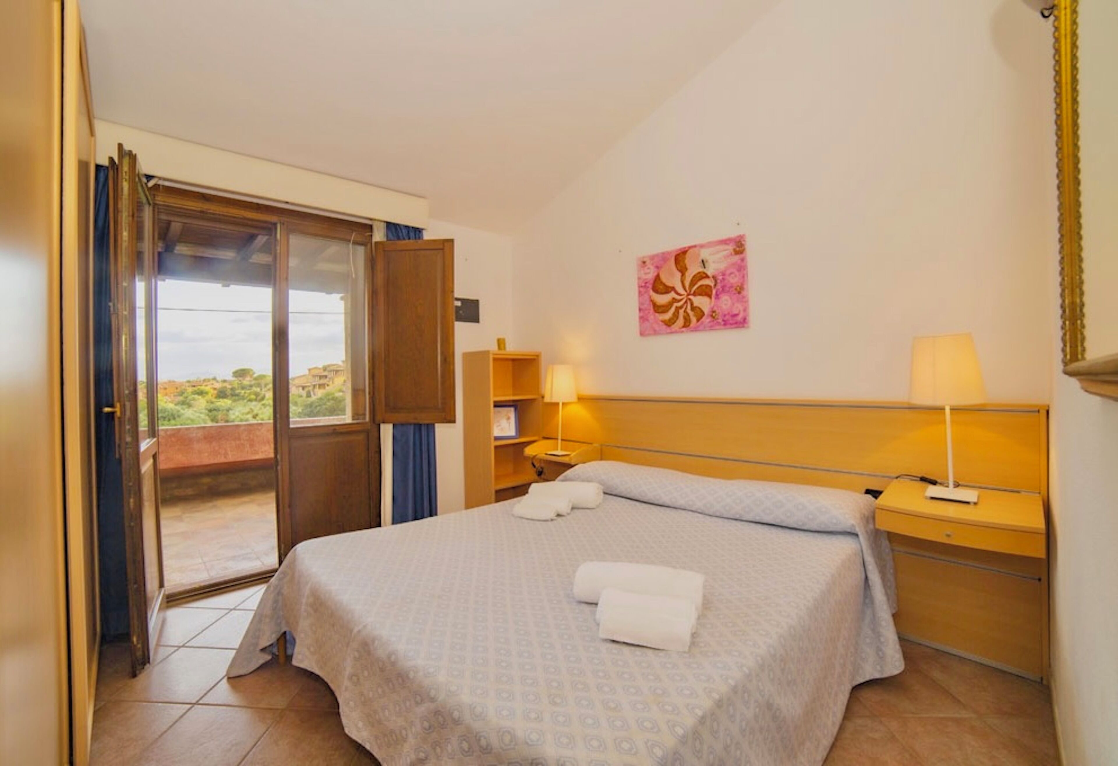 A hotel room with a bed, two bedside tables, a window with a view, a door, and a painting on the wall.
