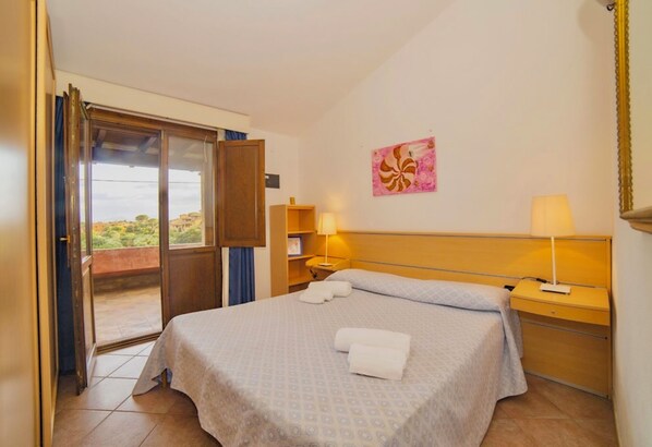 A hotel room with a bed, two bedside tables, a window with a view, a door, and a painting on the wall. - Village in Porto Corallo  (Villaputzu)