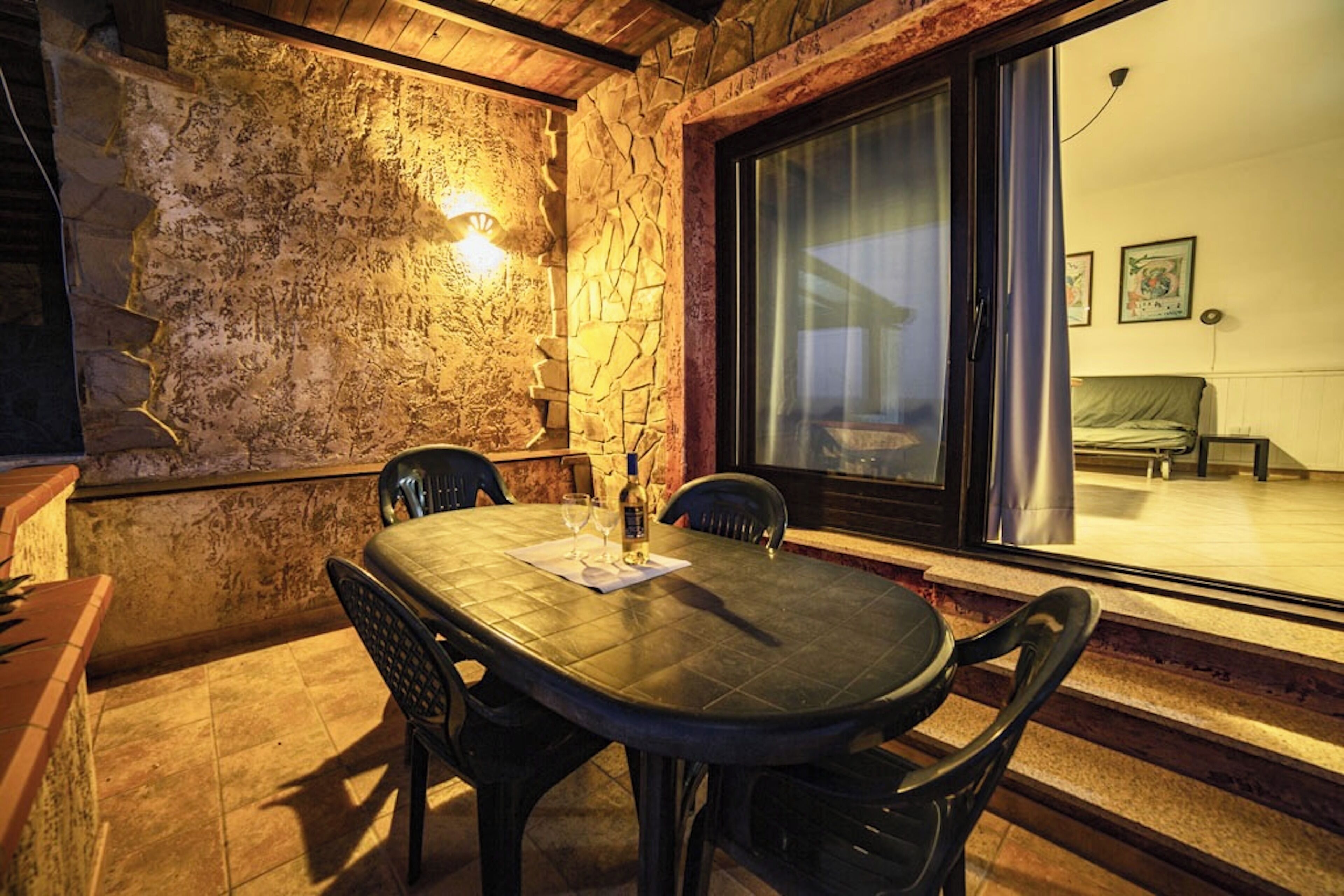 A stone-walled dining area with a table and chairs, a lit wall-mounted light, and a view through a glass door to an interior room with a bed and a chair.